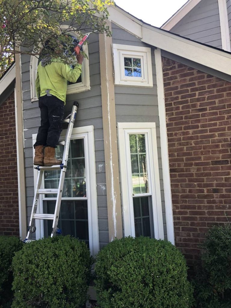 Painter on a ladder painting gray siding and white window trim